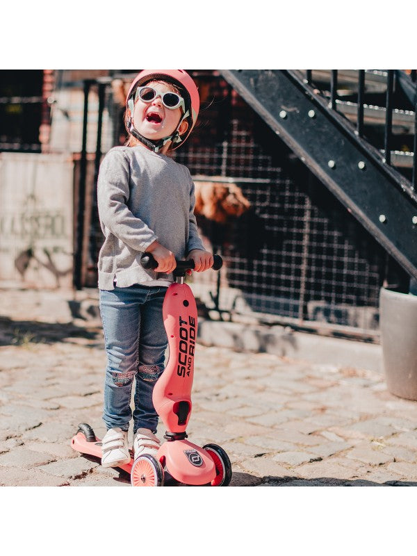 Kind op de Scoot and Ride - Highwaykick 1 - Peach met een helm en zonnebril, lachend op een stenen pad.